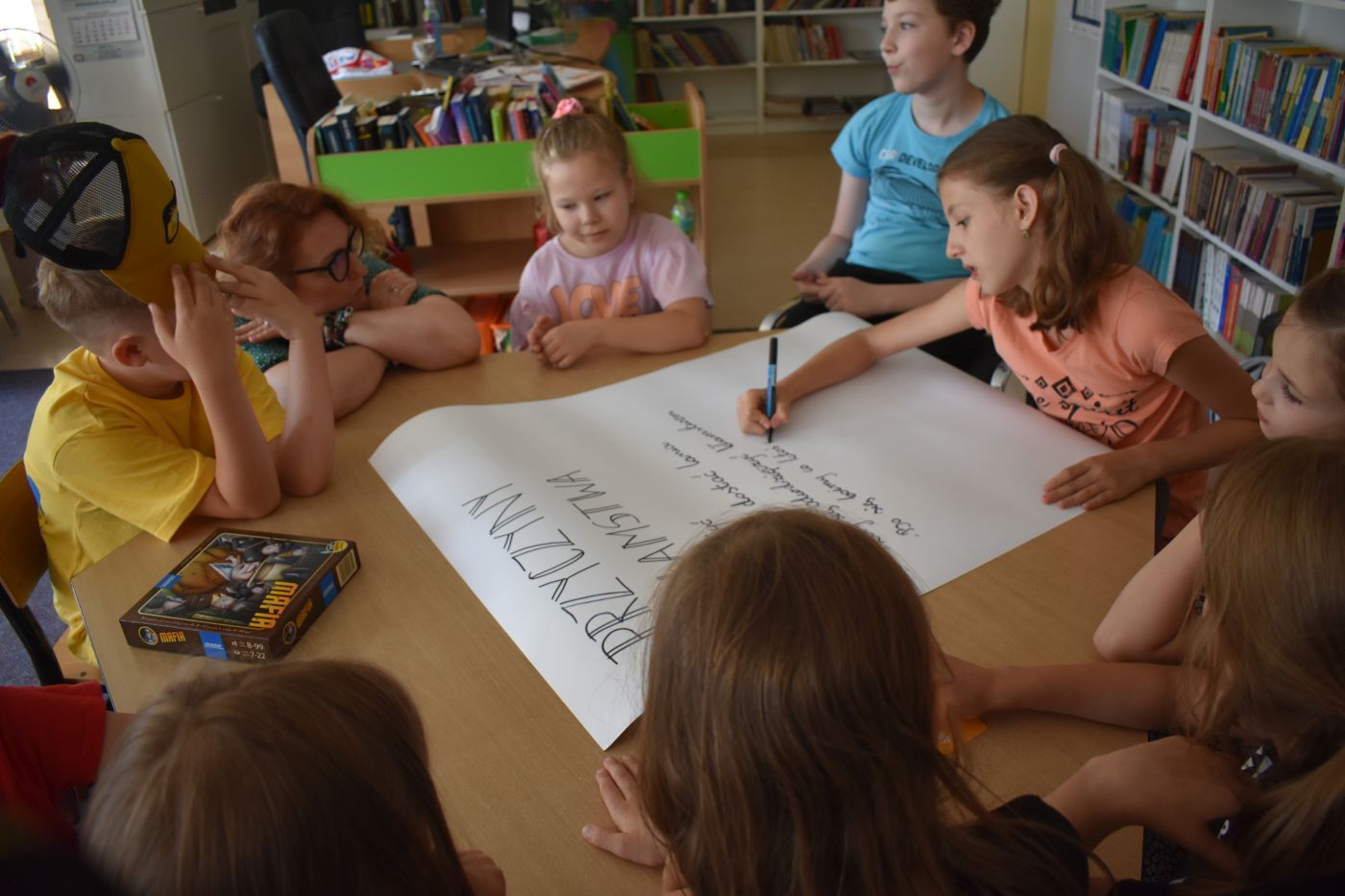 Dzieci przy stoliku rozmawiają a dziewczynka pisze na dużej kartce